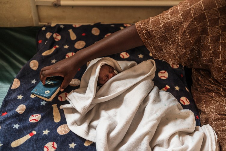 A photograph of an infant wrapped in blankets, lying on a bed with baseball-theme sheets. A woman’s hand reaches over the baby to pick up the Bible lying on the bed.