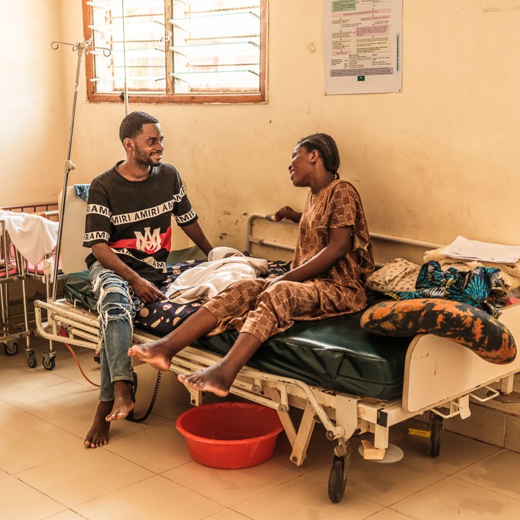A photograph of a man and a woman sitting on a hospital bed smiling at each other. A blanket-wrapped infant lies on the bed between them.