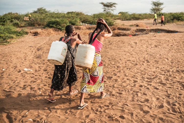 Two women walk in a desert setting, on brown sand with patches of green scrub brush, carrying jerricans on their backs.