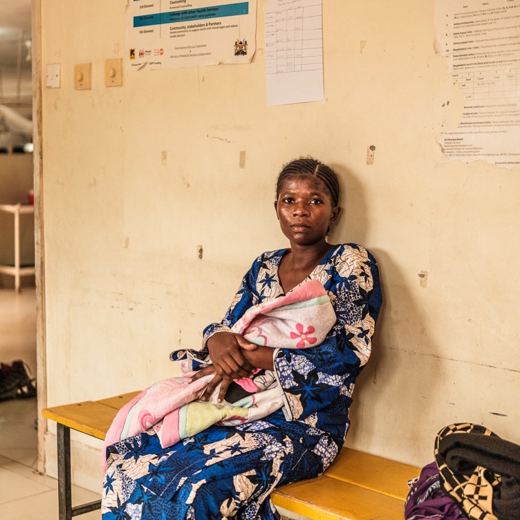 A photograph of a woman holding a blanket-wrapped infant, looking at the camera. She sits on a bench in a simple hospital room, with yellow walls covered in scratches and pieces of tape.