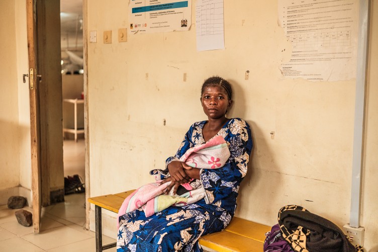 A photograph of a woman holding a blanket-wrapped infant, looking at the camera. She sits on a bench in a simple hospital room, with yellow walls covered in scratches and pieces of tape.