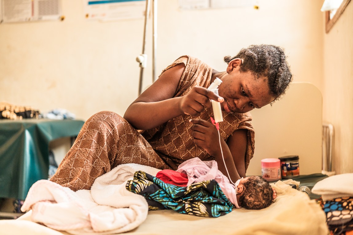 A photograph of a woman holding a device with a tube leading down to the mouth of a tiny, malnourished infant.
