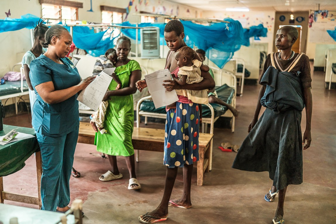 Several women hold babies and paperwork as they wait to speak with a woman in a nurse’s uniform, in a room filled with hospital beds, each with a bright blue mosquito net above it.