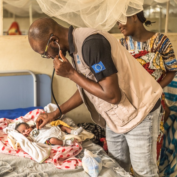 A photograph of a man leaning over an infant, listening to the child’s chest with a stethoscope. A woman stands behind him and looks on. They are in a hospital room with numerous hospital beds made up of simple metal frames and thin mattresses, each with a mosquito net hanging overhead.