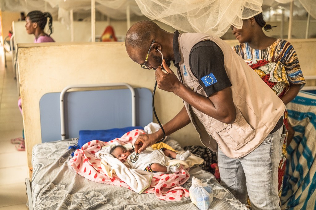 A photograph of a man leaning over an infant, listening to the child’s chest with a stethoscope. A woman stands behind him and looks on. They are in a hospital room with numerous hospital beds made up of simple metal frames and thin mattresses, each with a mosquito net hanging overhead.