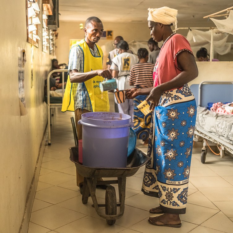 A photograph of a man pouring porridge into a mug that a woman is holding. The two of them are in a hospital room, with simple beds and mosquito nets in the background. The porridge is in a large purple barrel, resting in a wheelbarrow.