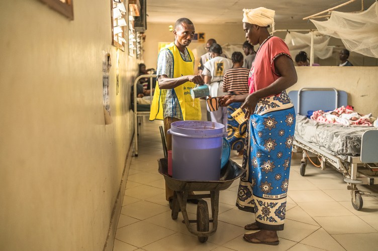 A photograph of a man pouring porridge into a mug that a woman is holding. The two of them are in a hospital room, with simple beds and mosquito nets in the background. The porridge is in a large purple barrel, resting in a wheelbarrow.