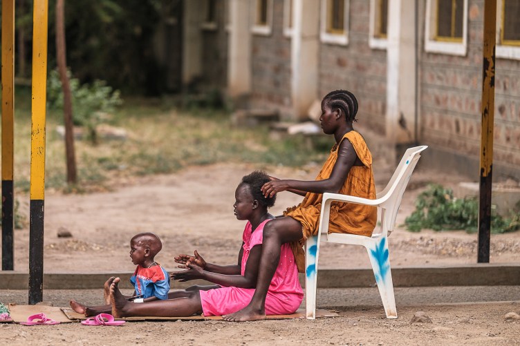 A woman with cornrows sits outdoors in a plastic chair, her hands in the hair of another woman sitting on the ground. The woman on the ground has her legs outstretched, and between them sits a small child.