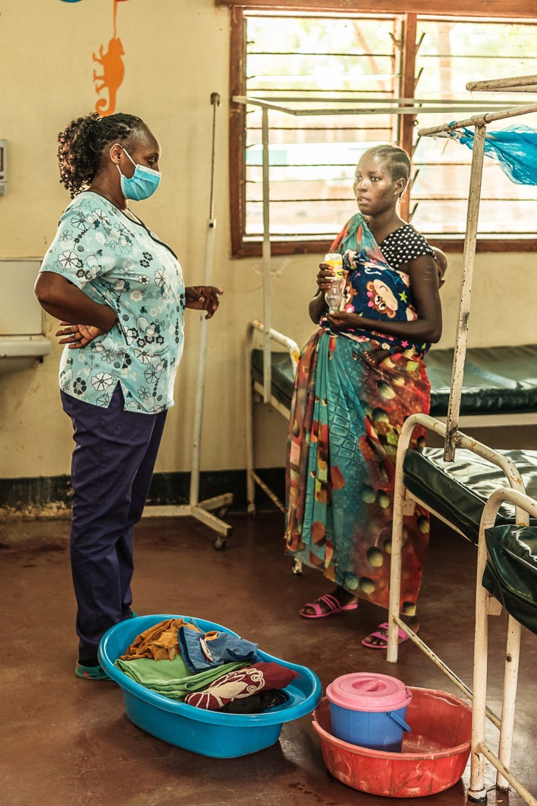 In a hospital room with rows of dilapidated metal beds, a woman with cornrows wearing a patterned wrap speaks with a nurse in hospital scrubs who has a curly ponytail and wears a mask. Two plastic tubs of belongings sit at their feet.