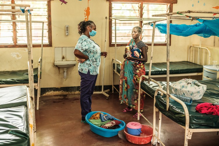 In a hospital room with rows of dilapidated metal beds, a woman with cornrows wearing a patterned wrap speaks with a nurse in hospital scrubs who has a curly ponytail and wears a mask. Two plastic tubs of belongings sit at their feet.