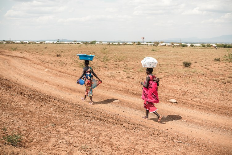 Two women in patterned wrap dresses walk along a dirt road in a stretch of desert. Each balances a tub of belongings on their head and carries a child tied to their back.