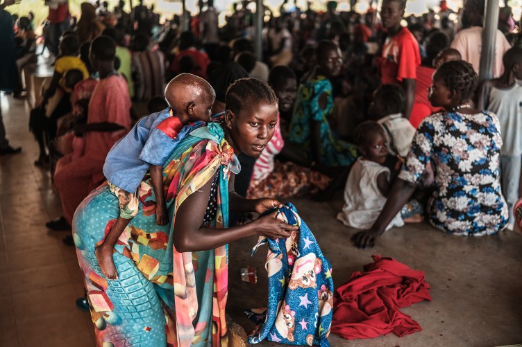 A woman in a patterned wrap leans over, her small child resting on her back as she prepares to tie him securely to her. Behind her, a crowd of other people, mostly women and children, wait.