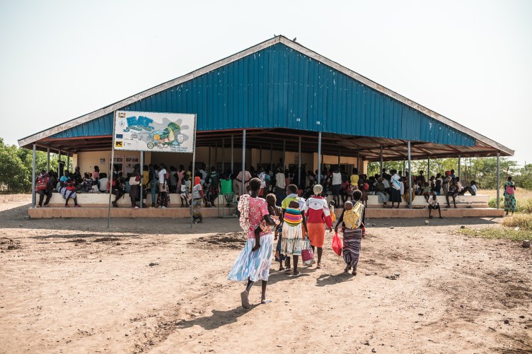 Several women holding children approach a large pavilion with long stone benches, where a crowd of people are already waiting.