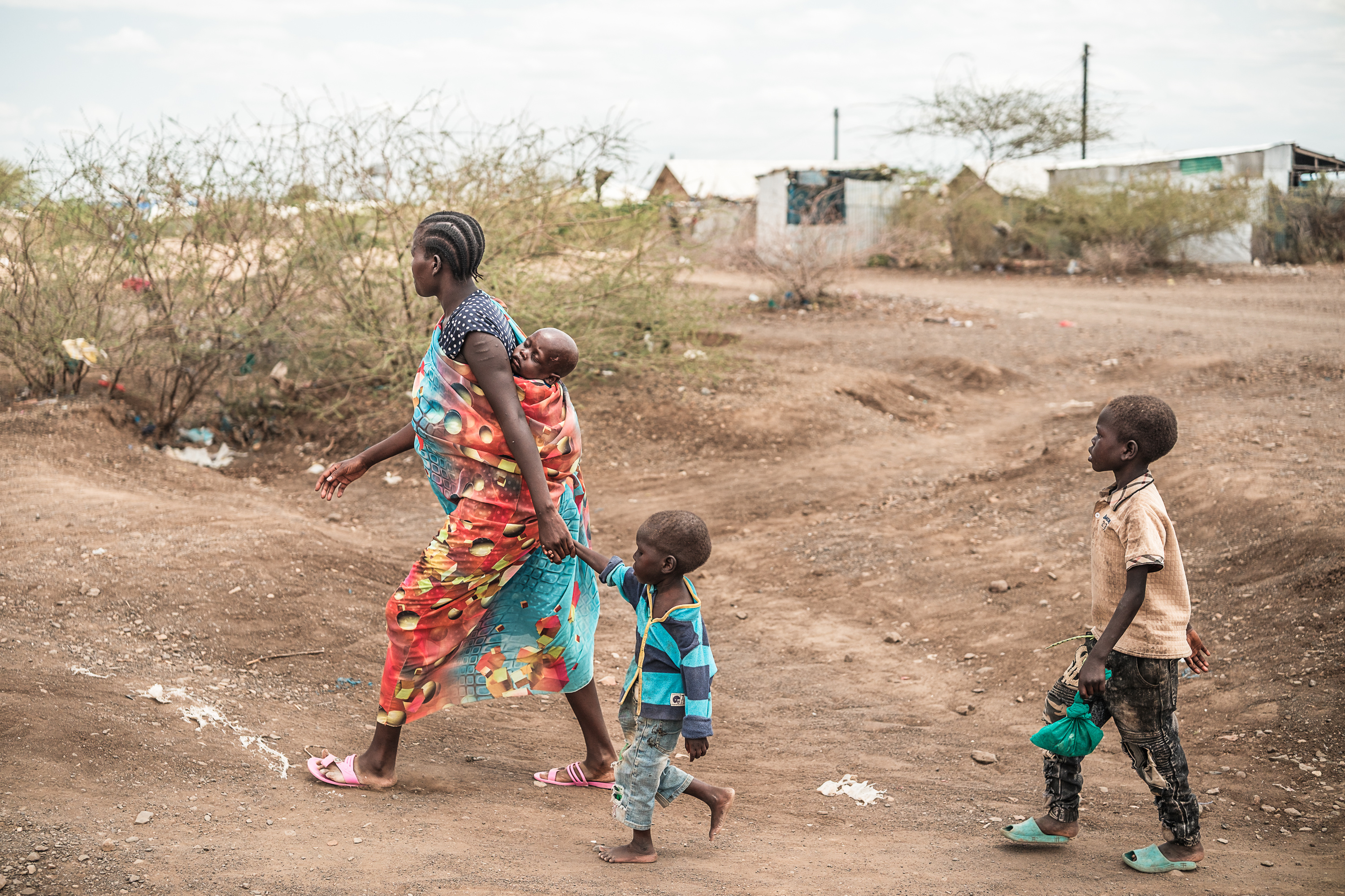 A woman in a patterned wrap dress strides along a dirt path with scrub brush and small houses in the background. She carries one small child tied to her back and holds another by the hand. A slightly taller child trails behind them.