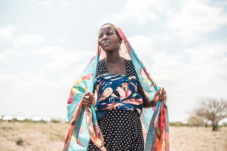 A woman in a polka-dot dress looks away from the camera as she drapes a patterned scarf on her head, framing her face.