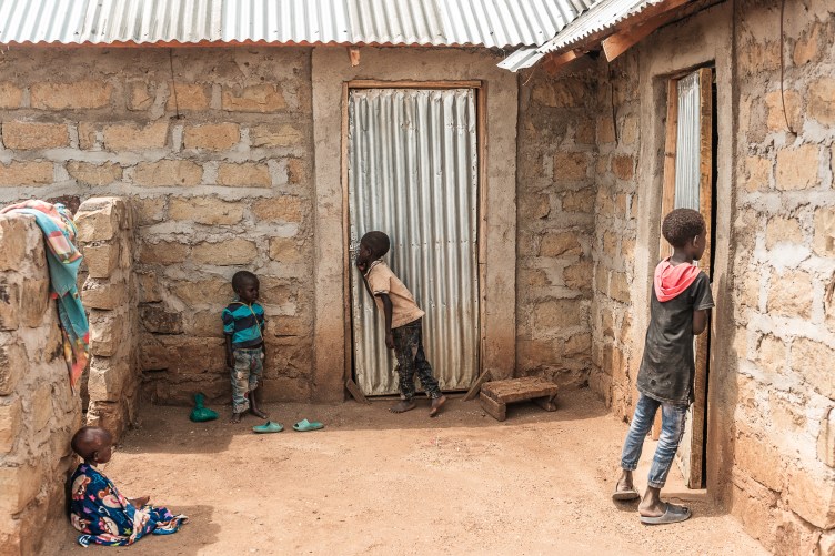 Against a backdrop of a simple brick house with a corrugated metal roof and doors, four small children wait. The smallest sits to the left in the shade in a patterned wrap. Another stands barefoot to his right, while a taller child beside him tries to peer into one door. The tallest child, on the far right, peers through a crack in another door.