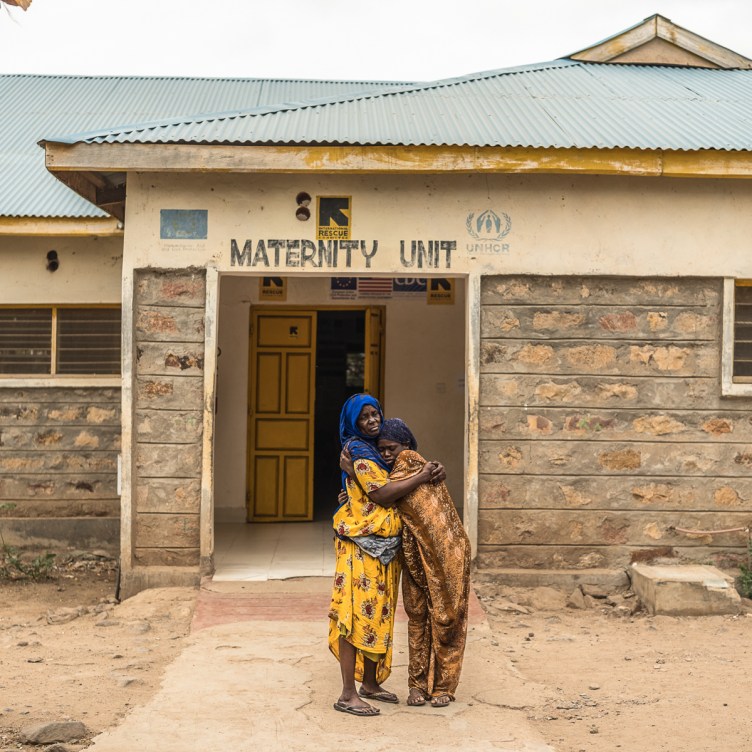A photograph of two women holding each other in front of a building that says Maternity Unit in faded letters. They are on a crumbling sidewalk in the midst of dirt. The building has visible cracks and fading paint.