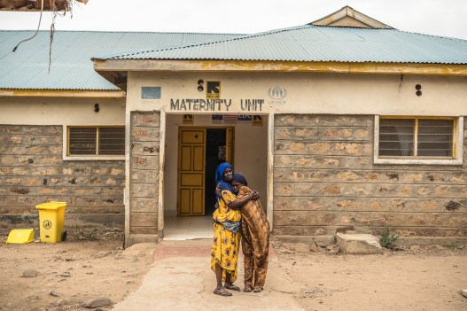 A photograph of two women holding each other in front of a building that says Maternity Unit in faded letters. They are on a crumbling sidewalk in the midst of dirt. The building has visible cracks and fading paint.