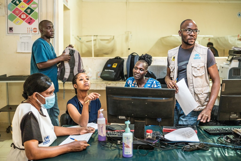 A photograph of several people hovering around a computer monitor and paperwork in a hospital setting. One of them stands as if to leave.
