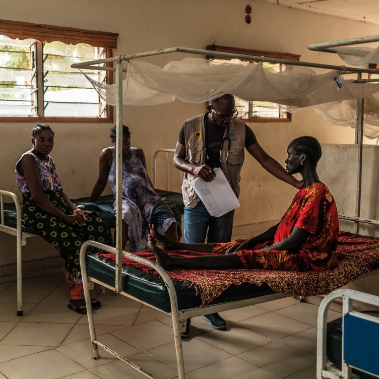 A photograph of a man with a stethoscope around his neck reaching his arm out to the shoulder of a woman sitting on a hospital bed.