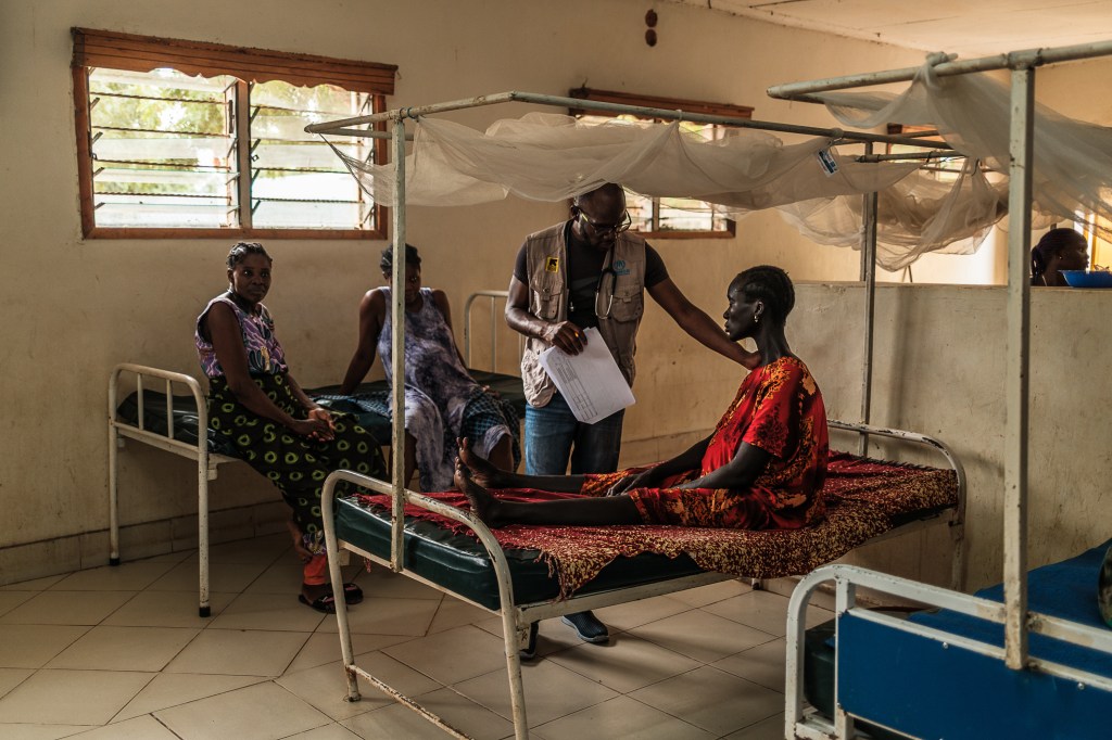 A photograph of a man with a stethoscope around his neck reaching his arm out to the shoulder of a woman sitting on a hospital bed.