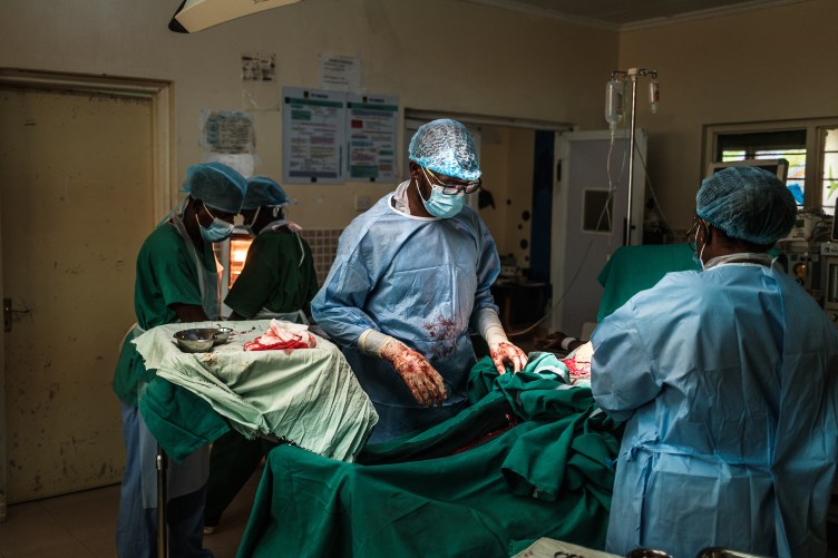 A photograph of a dark hospital room in which several people wearing surgical gowns, caps, gloves and masks hover over a woman who is completely covered in surgical sheets. The hands of the person in the center are covered in blood.