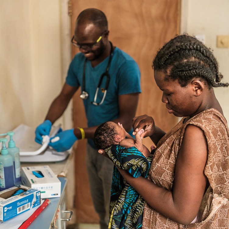 A woman cradles a tiny infant in one hand while holding the baby’s hand with her other hand. They are in a hospital room. In the background, a man with a stethoscope around his neck prepares medical equipment.