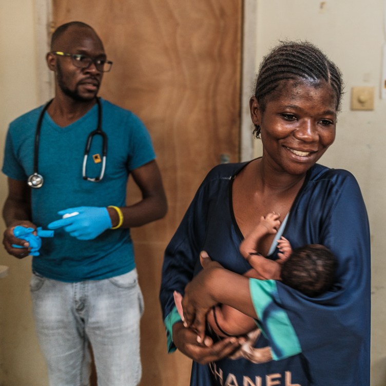 A woman smiles as she holds a small infant. Behind her, a man with a stethoscope and medical gloves has a concerned look on his face.