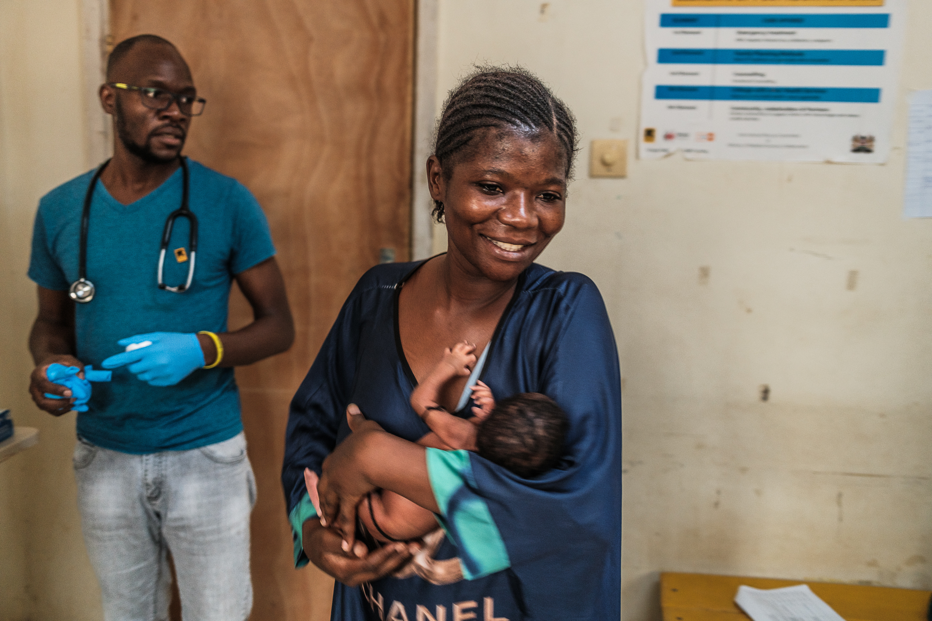 A woman smiles as she holds a small infant. Behind her, a man with a stethoscope and medical gloves has a concerned look on his face.
