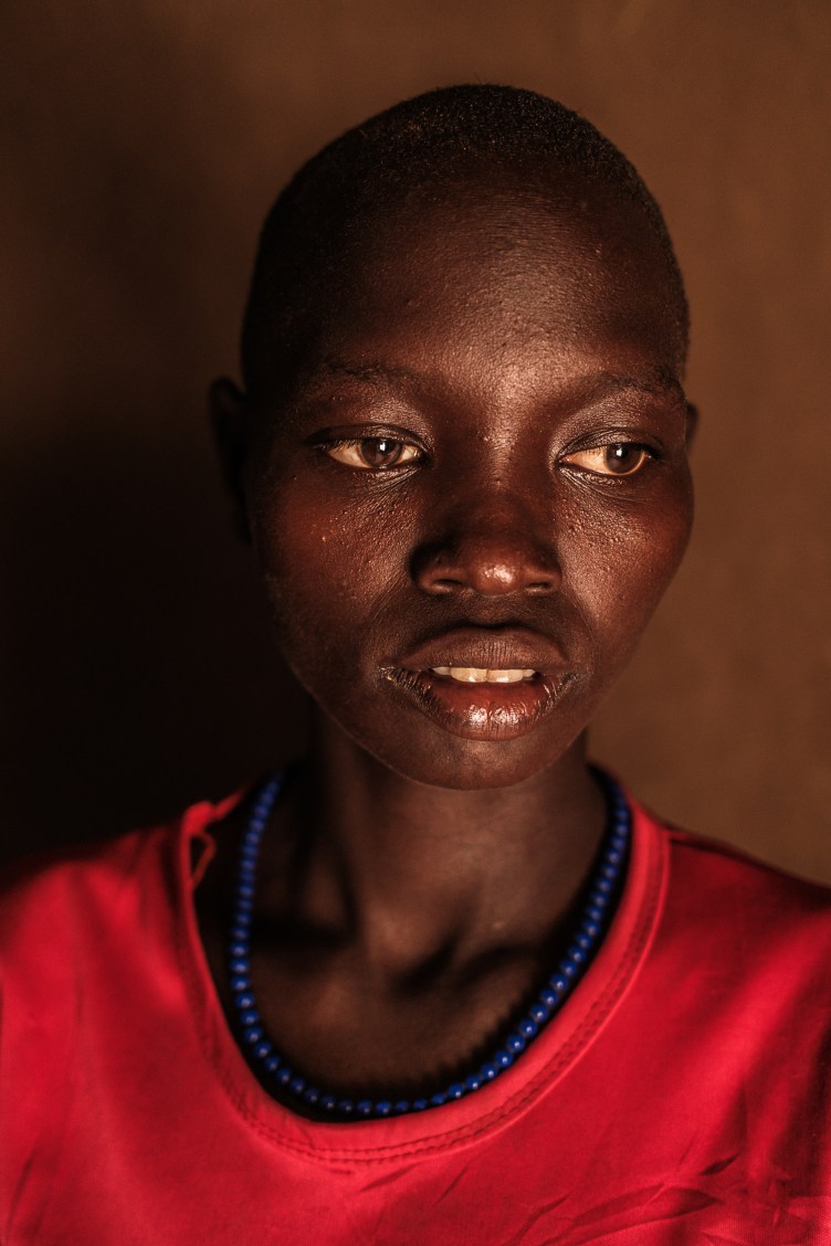 A headshot of a young woman, who looks off to the side. She is wearing a red T-shirt and a blue necklace.