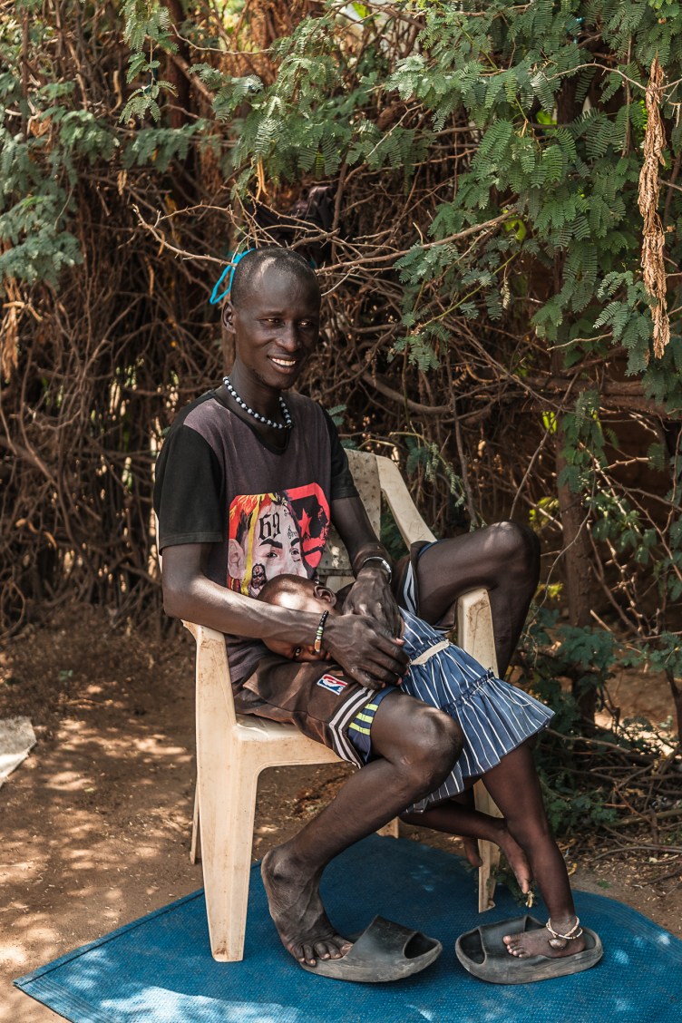 A man sits on a plastic chair. A small girl stands next to him, resting her head in his lap as he holds her. They are outdoors, with a dirt floor, surrounded by scrub brush and simple fencing.