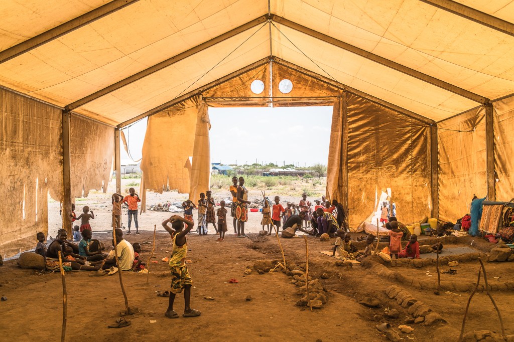 Under a large brown tent, people mill about, playing, conversing, and resting. Rocks and sticks on the ground mark sleeping areas for people and families.