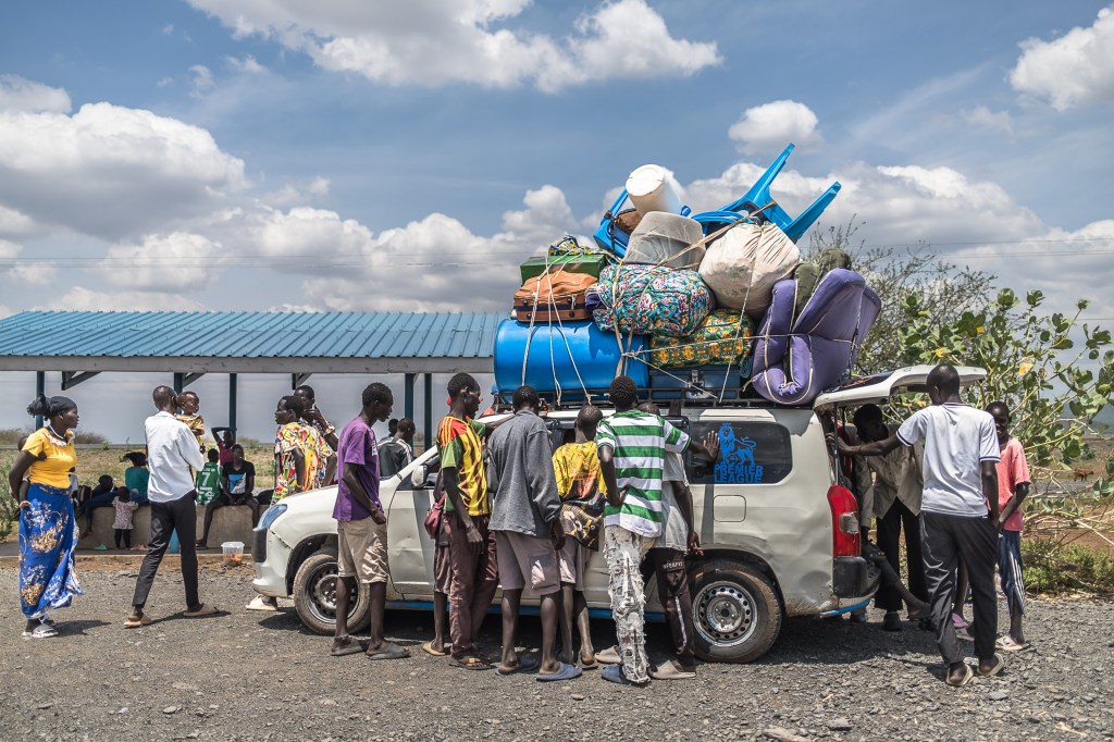 People gather around a hatchback car. Several of them are climbing in the back even though the car is already full. Luggage and furniture are piled high on top of the car and tied in place.