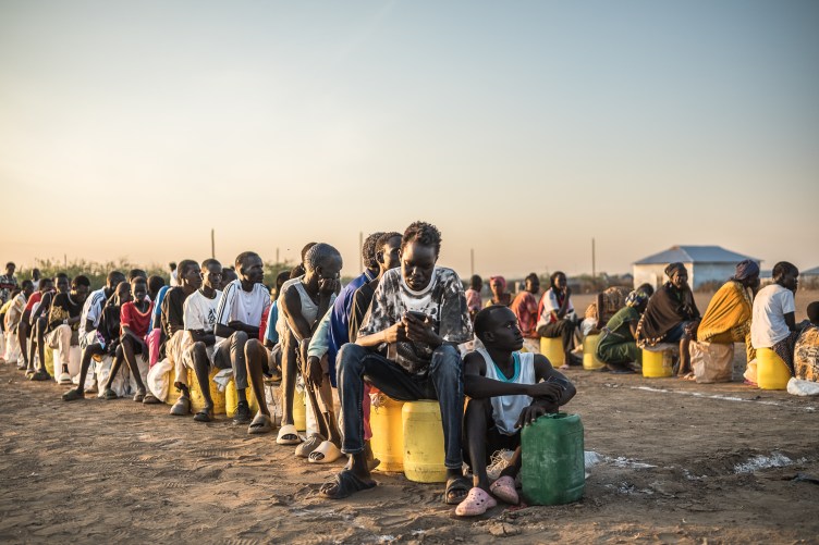 A photograph of dozens of people sitting, on jerricans or on the ground, in two rows.