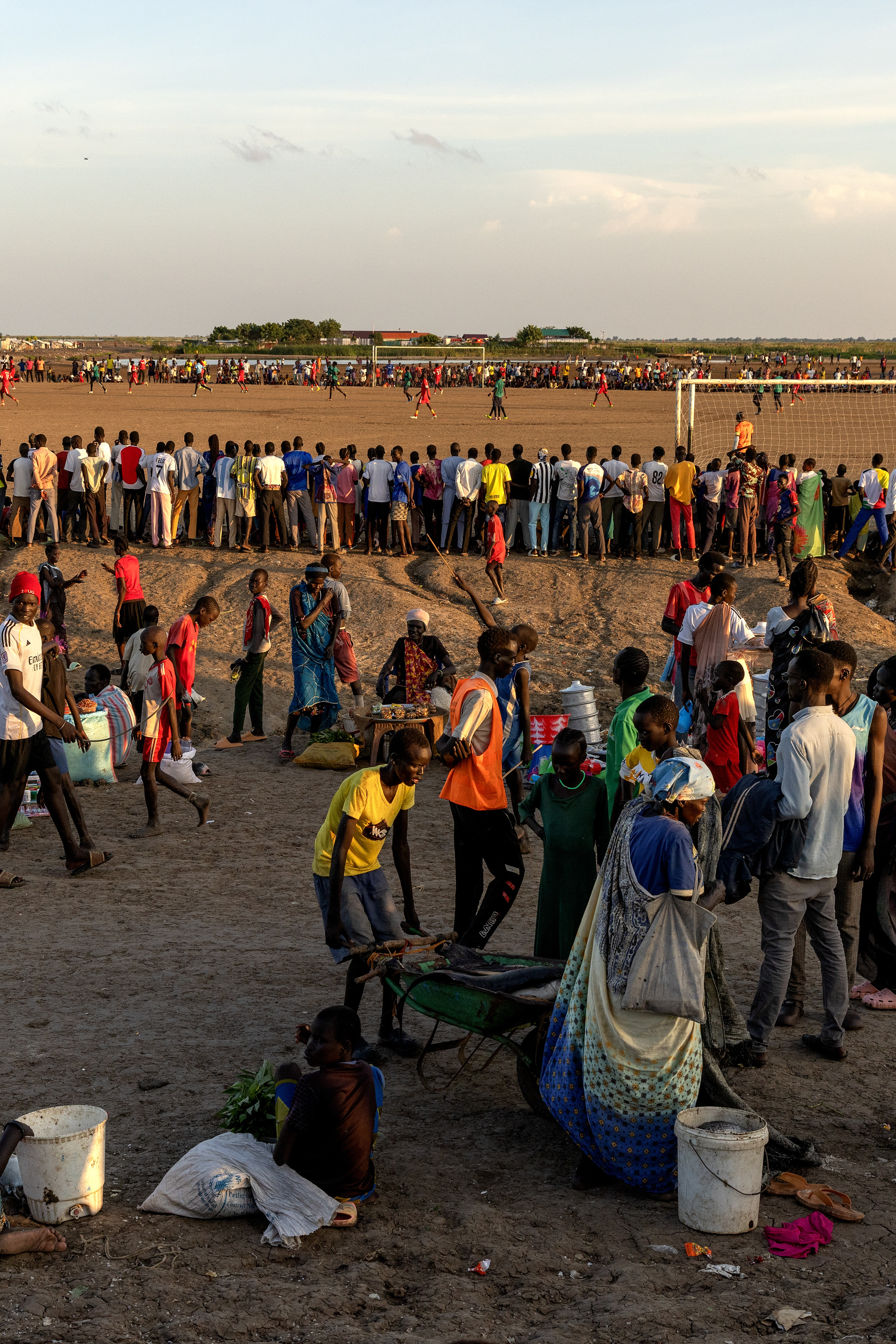 Hundreds of people gather on a large patch of dirt to watch people playing soccer. In the foreground, people walk on a dirt road, one of them pushing a wheelbarrow.