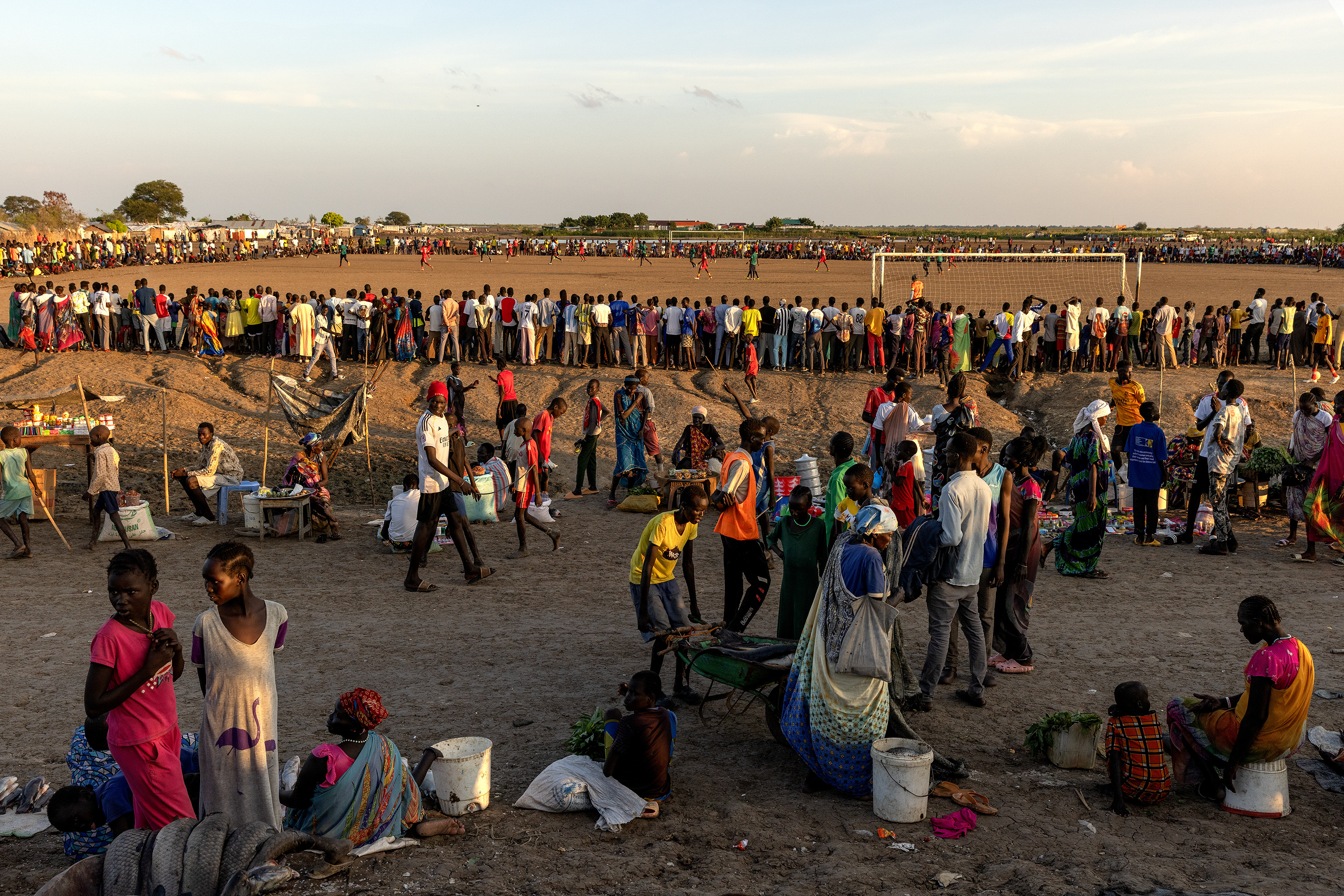 Hundreds of people gather on a large patch of dirt to watch people playing soccer. In the foreground, people walk on a dirt road, one of them pushing a wheelbarrow.