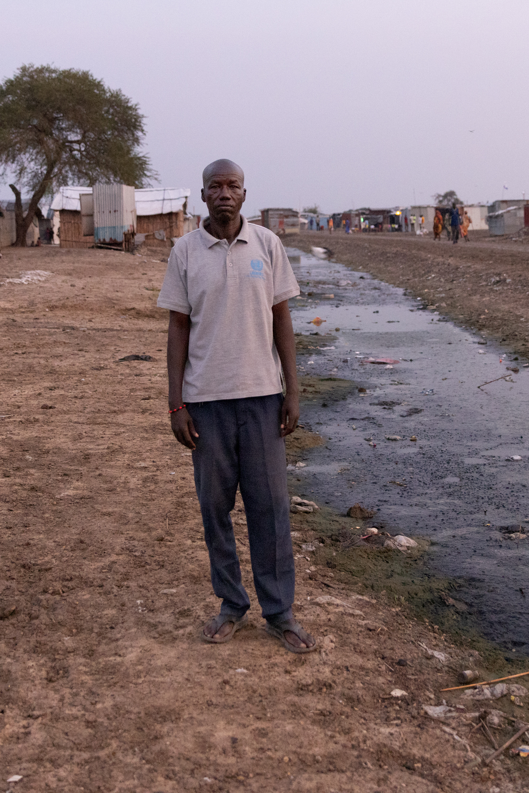 A man stands in the center of a refugee camp, next to a muddy culvert that is full of trash, feces and mud. In the background are people walking by and simple homes made from straw.