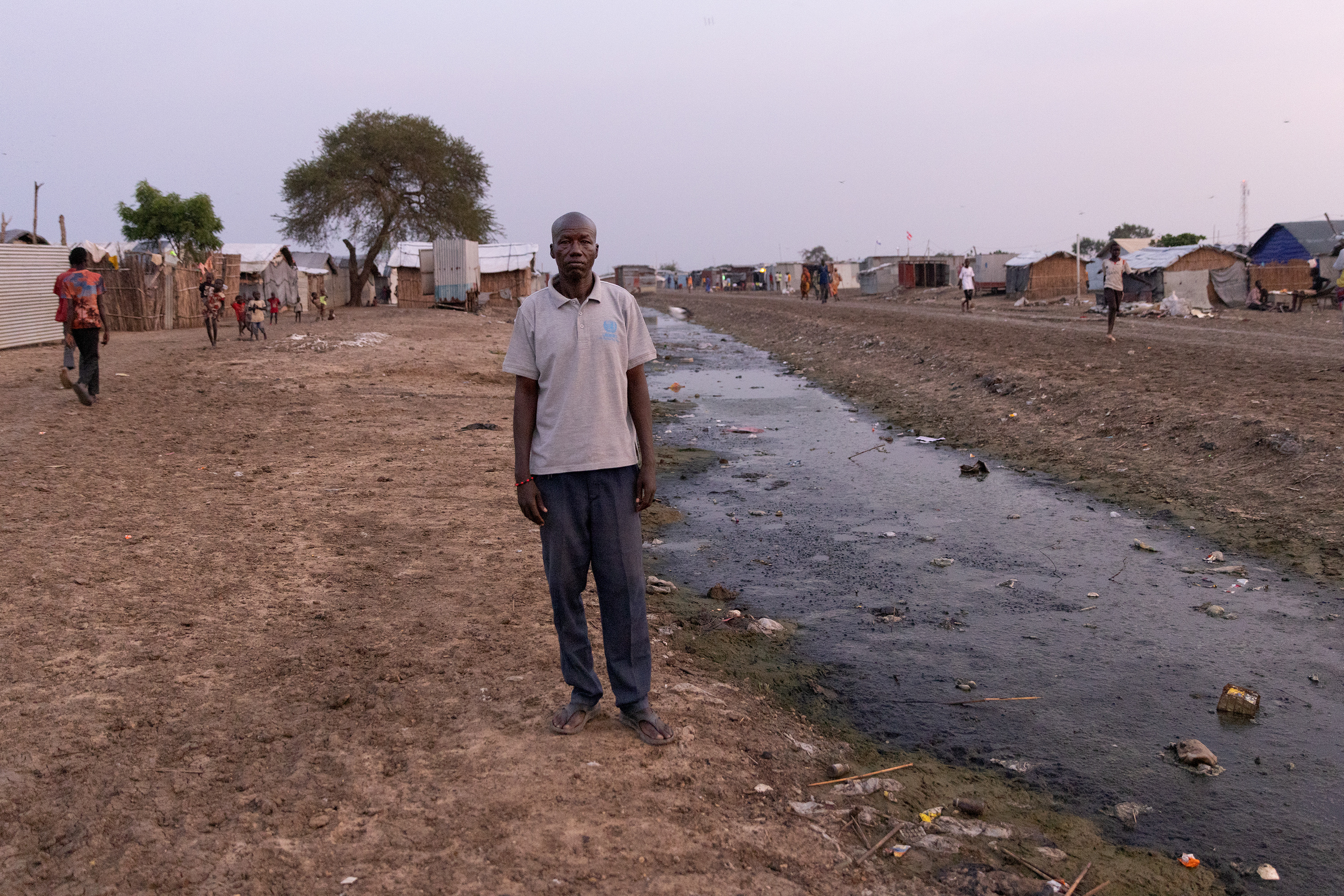 A man stands in the center of a refugee camp, next to a muddy culvert that is full of trash, feces and mud. In the background are people walking by and simple homes made from straw.