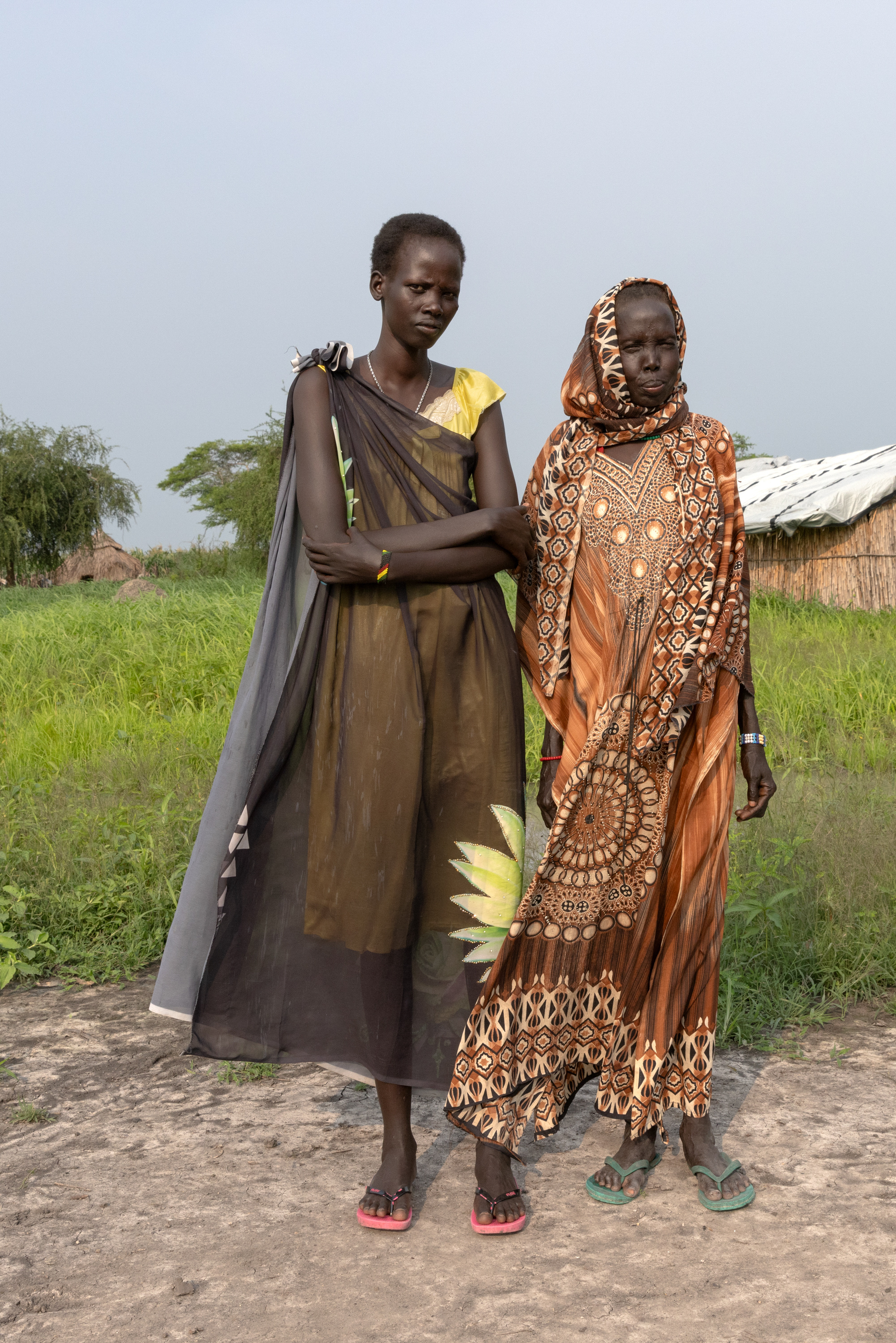 Two women stand and look at the camera wearing brightly colored clothes in front of a field, trees and several homes made of woven straw.