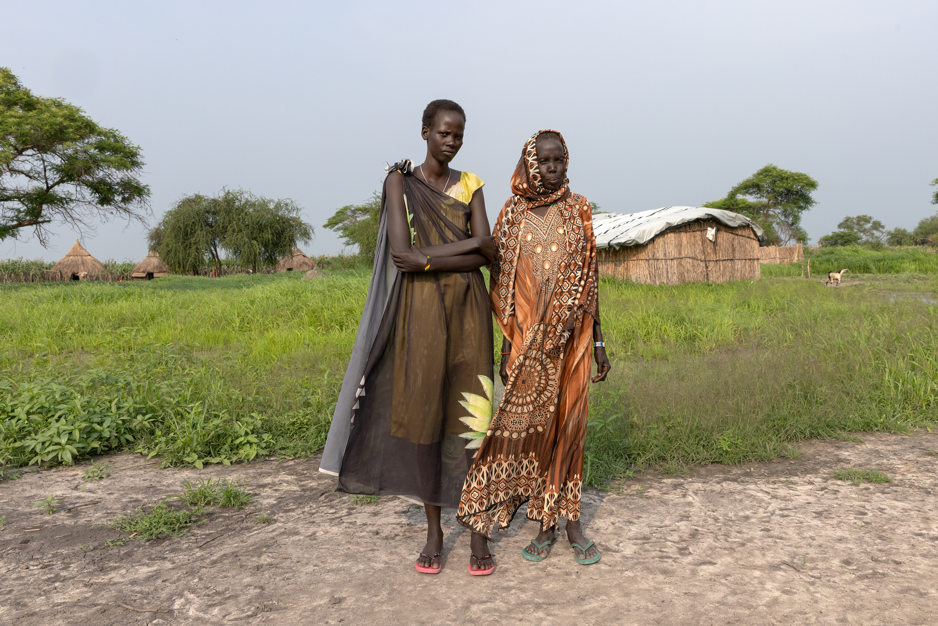Two women stand and look at the camera wearing brightly colored clothes in front of a field, trees and several homes made of woven straw.