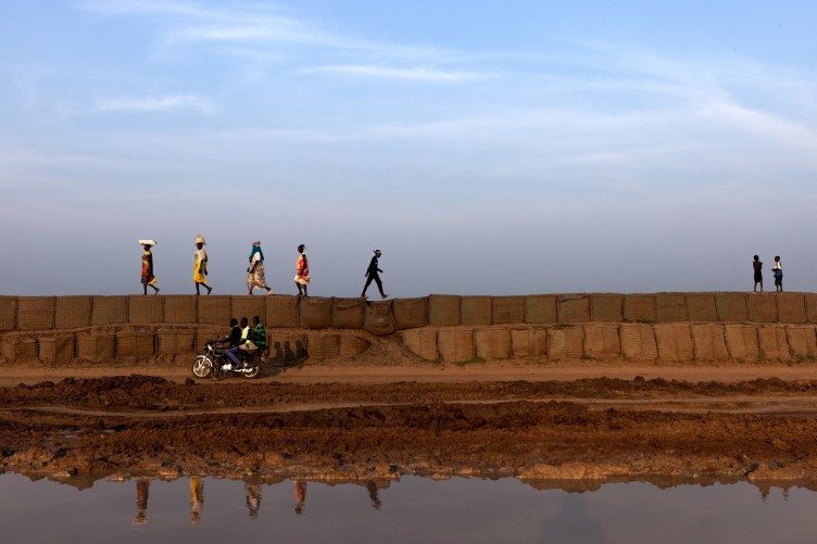People travel on a muddy dirt road that is partially washed away. A motorbike carries three people on part of the road. People walk on barriers meant to hold back floodwater. They are silhouetted against the sky, and their reflections appear in the water below.