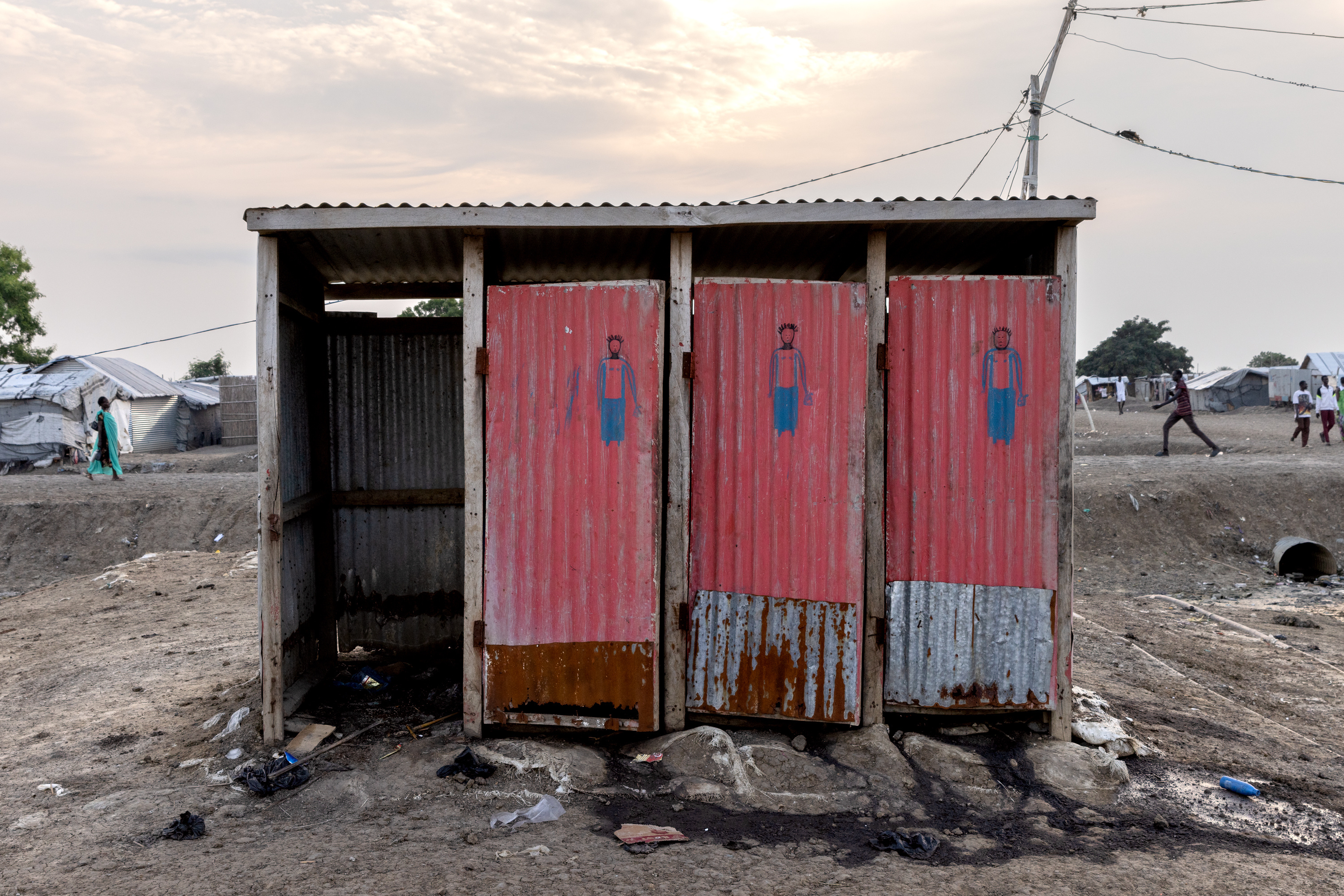 A latrine in the midst of a refugee camp. One of the stalls is missing a door and is overflowing, and the remaining stalls all have liquid flowing from them into a nearby culvert.
