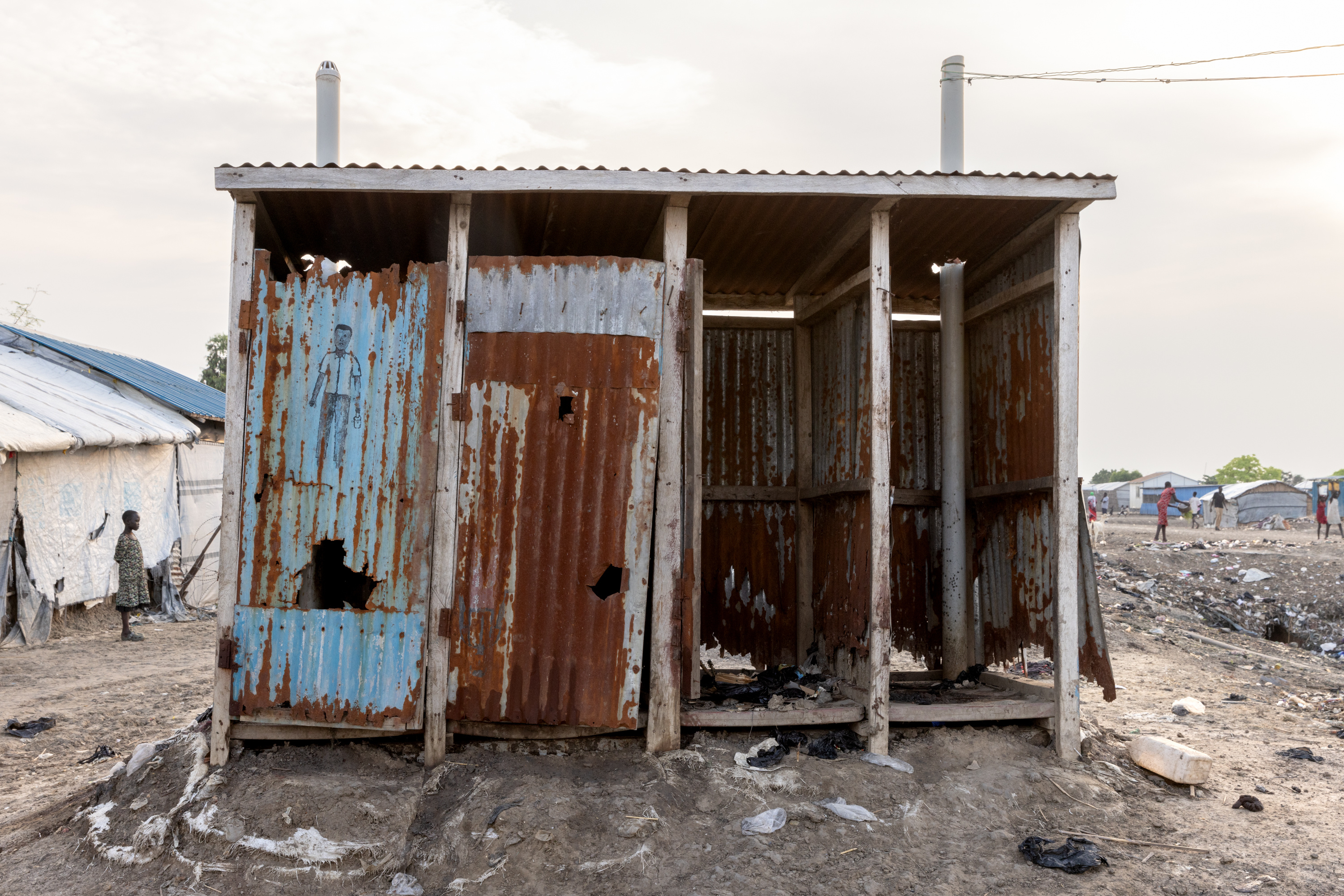 A latrine in the midst of a refugee camp. The latrine is dilapidated, with two missing doors and numerous holes in the walls.
