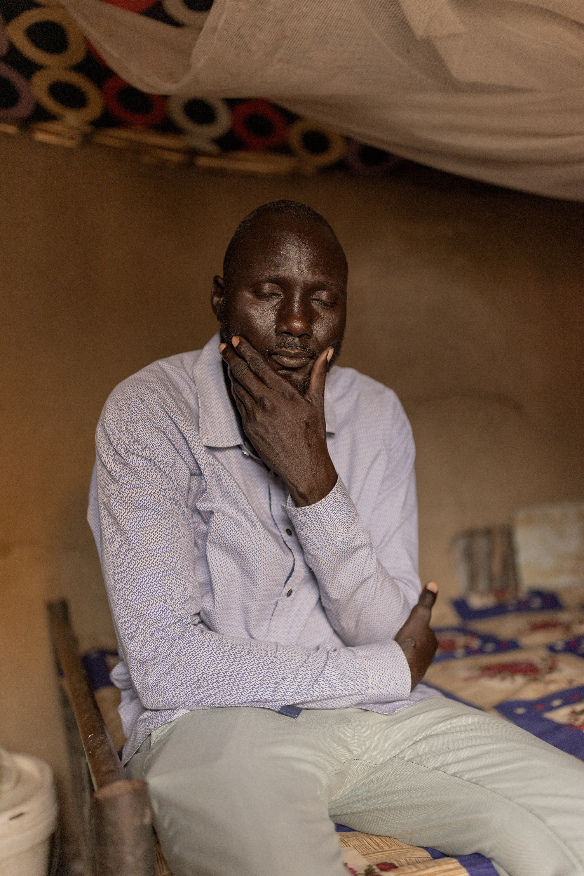 A man sits on a bed, with a mosquito net hanging above him, in a room with brown mud walls. He has his eyes closed and is holding his chin with one hand.