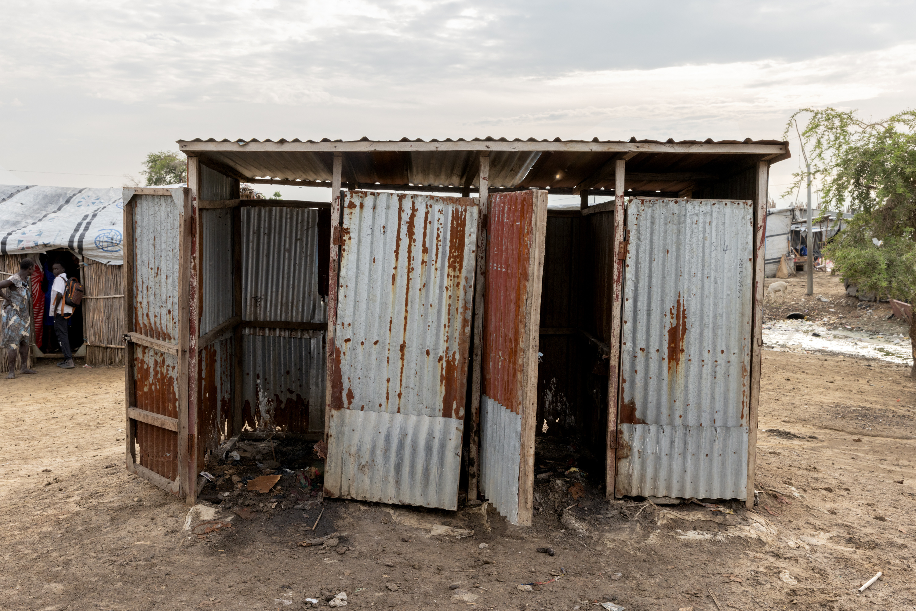 A latrine in the midst of a refugee camp with overflowing stalls and holes in the walls.