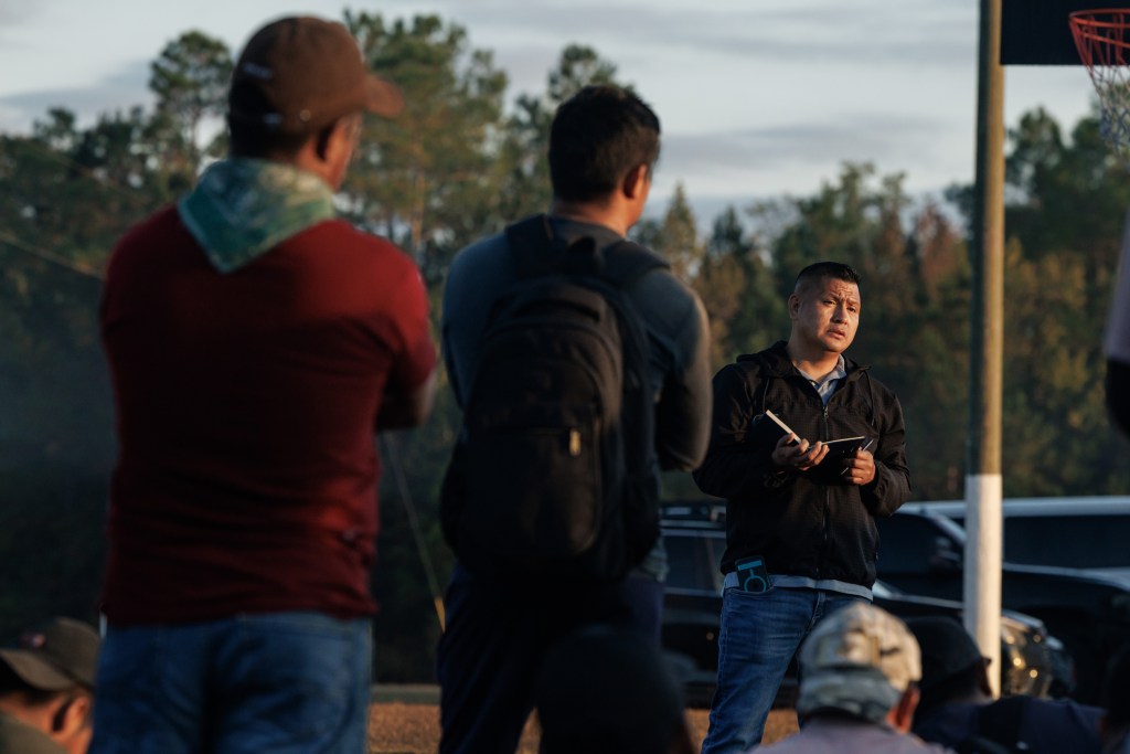 A group of men who are standing and sitting listen to Salucio as he holds an open book. They are outside on a basketball court with trees in the distance.