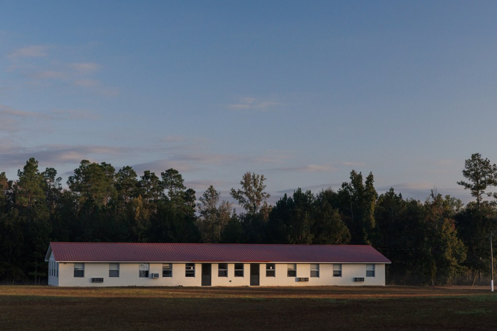 A one-story building with two doors and windows all along its face sits in front of a line of trees. Above are a blue sky and clouds.