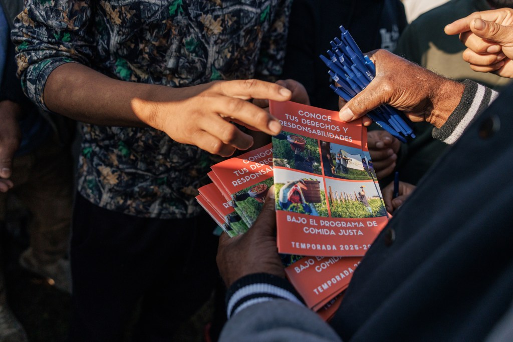 Someone holds pens and a stack of literature with the Spanish words “Conoce tus derechos y responsabilidades bajo el programa de comida justa.” Other people’s hands reach for the pamphlets.