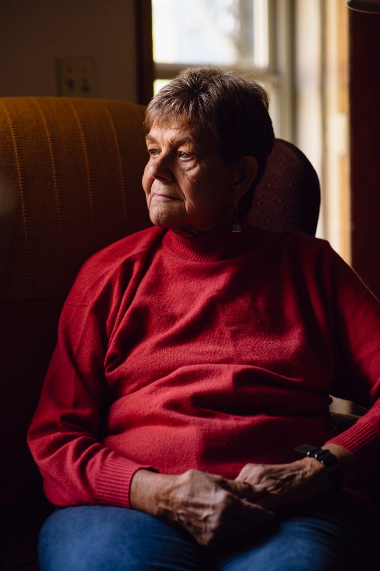 A woman sits in a living room setting, looking to the side, with her hands clasped in her lap.