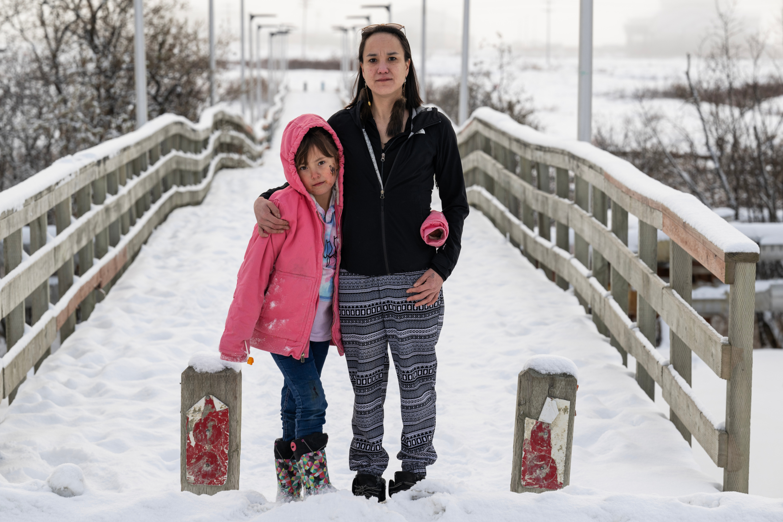 A woman wearing long fur earrings, a black jacket and patterned pants stands on a snow-covered boardwalk with a small girl wearing a pink jacket, multicolored boots and a tie-dyed sweatshirt. The girl has a cat sticker on her cheek.